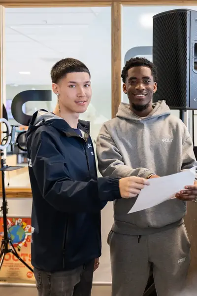Twee jonge mannen staan met een papiertje bij DJ-apparatuur in een creatieve ruimte met posters en speakers.