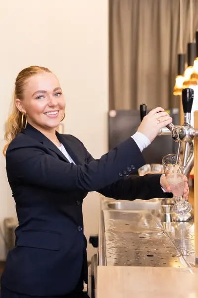 Vrouwelijke hotelmedewerker in donker pak tapt een drankje achter de bar.