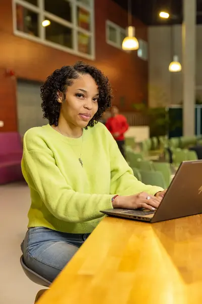 Student met laptop werkt in de kantine met houten muren en hanglampen, zittend aan een tafel.