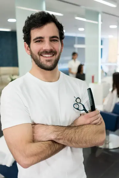 Een kapper met een schaar en kam in zijn hand staat in een praktijklokaal met op de achtergrond meerdere kappersstoelen waar studenten aan het knippen zijn.  