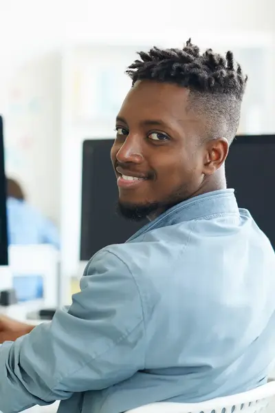 Man met een blouse aan zit achter achter een groot scherm te coderen. Hij kijkt over zijn schouder lachend de camera in.  Op de achtergrond staat een tweede scherm.