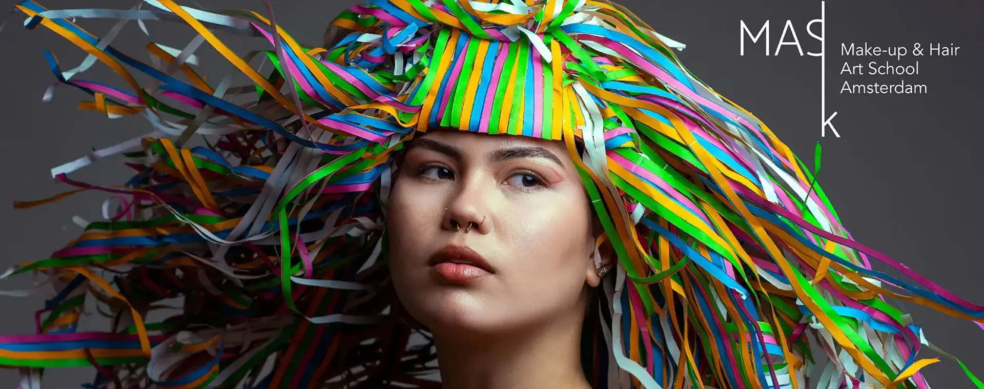 Model draagt kleurrijk hoofddeksel van linten, met creatieve make-up en neutrale achtergrond, voor Make-up & Hair Art School Amsterdam.
