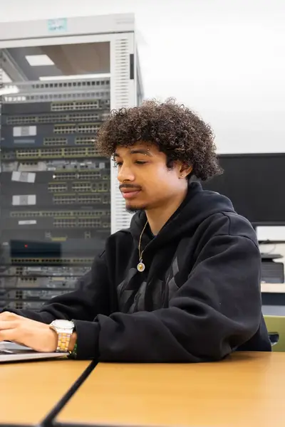 Student werkt op een laptop in een ICT-lokaal met netwerkapparatuur op de achtergrond.