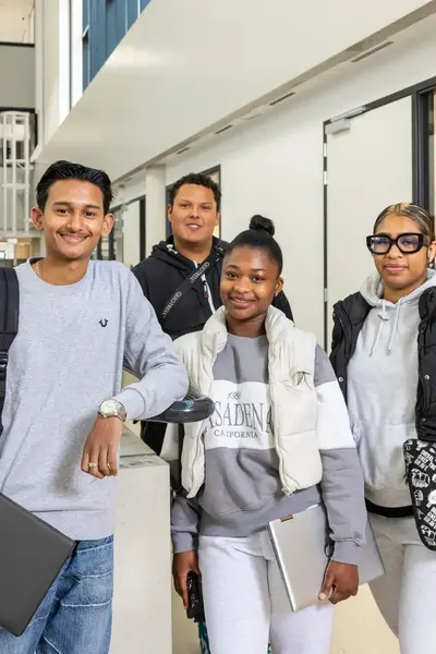 Vier studenten staan samen in de schoolgang en lachen naar de camera. Ze dragen laptops en rugzakken.