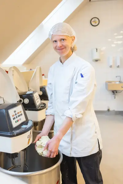 Student in bakkerskleding met haarnetje houdt deeg vast boven een grote deegmixer.