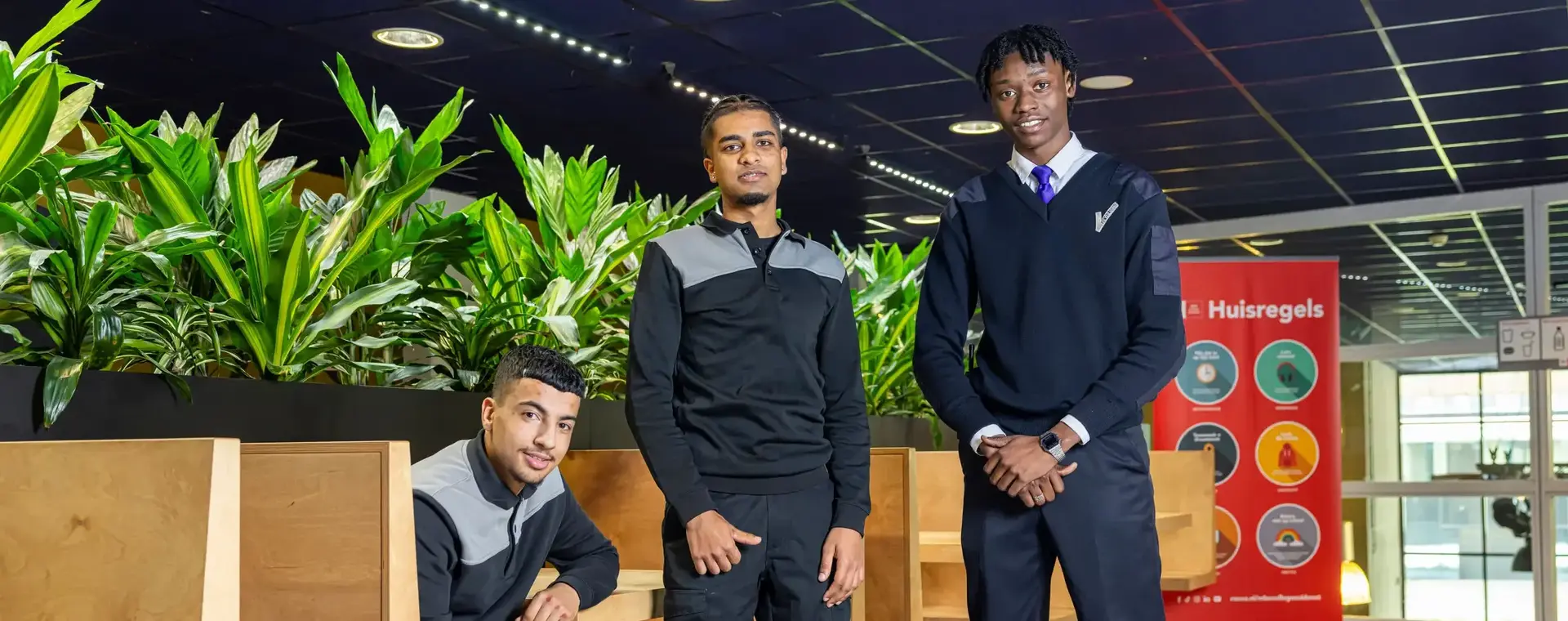 Drie studenten in beveiligingsuniform poseren in een schoolomgeving met groene planten en een bord met huisregels op de achtergrond.