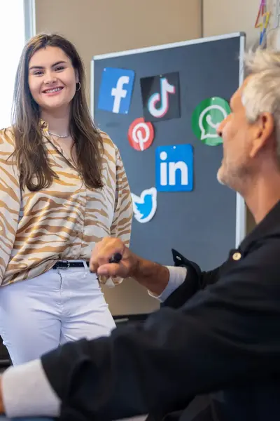 Student praat met docent in klaslokaal en twee studenten werkend op de achtergrond.