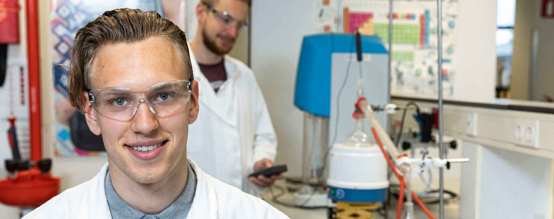 Twee studenten in laboratoriumjassen in een scheikundelokaal; één student glimlacht naar de camera, terwijl de ander op de achtergrond werkt met laboratoriumapparatuur.