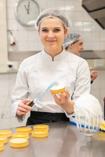 Een student patisserie glimlacht terwijl ze een taartbodem insmeert met crème. Ze draagt een witte bakkersjas en haarnetje.
