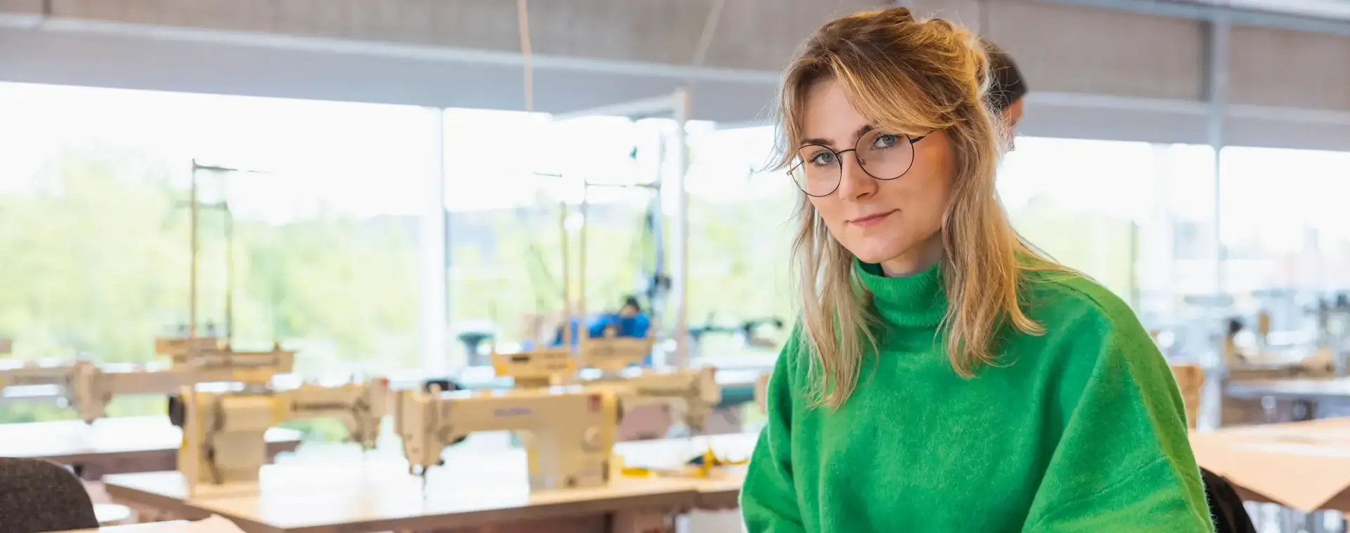 Jonge vrouw met groen trui aan achter tekentafel met op de achtergrond naaimachines.