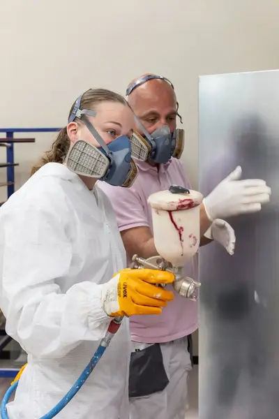Student in een overall met masker op is een paneel aan het bespuiten met witte verf. Een docent met masker op kijkt toe en geeft aanwijzingen.