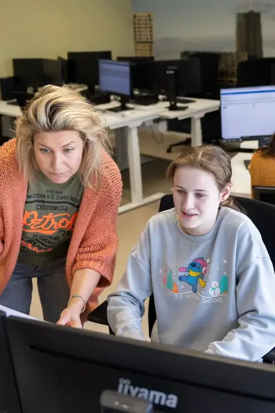 Studente achter computerschermen met docente naast haar die aanwijzingen geeft.