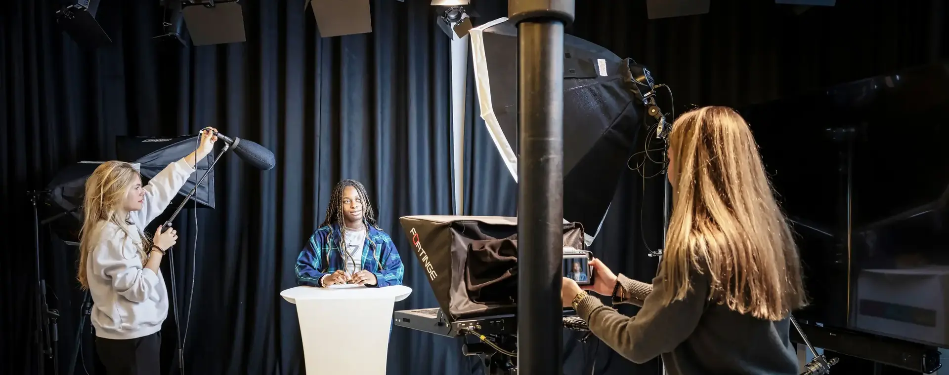 drie studenten staan in studio, eentje houdt een microfoon hoog vast, eentje is de presentator en eentje operates een camera