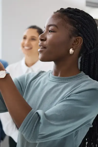 Een jonge vrouw schrijft iets op een whiteboard tijdens een presentatie, terwijl twee collega’s geïnteresseerd en glimlachend toekijken op de achtergrond.