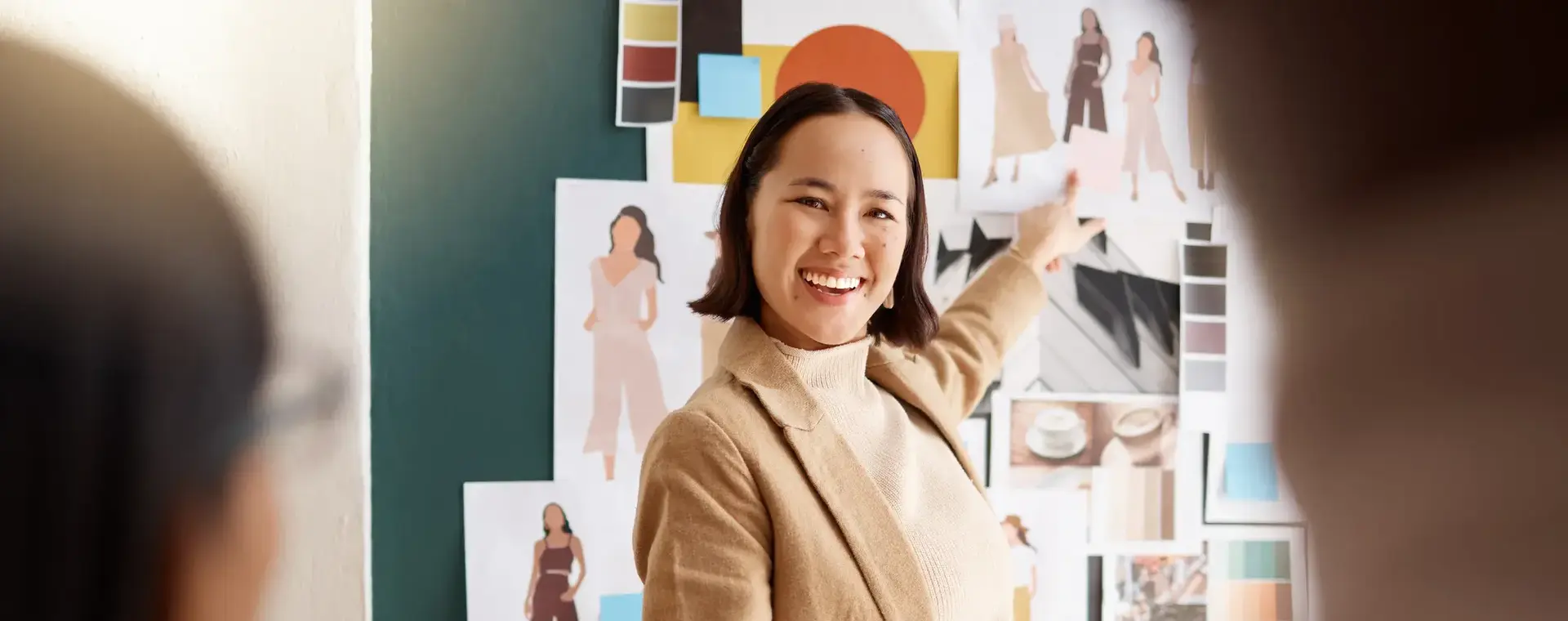 Een jonge vrouw staat met een map en pen in haar hand voor een mood board met allemaal designs van kleding. Ze wijst naar een tekening met verschillende kledingstukken.