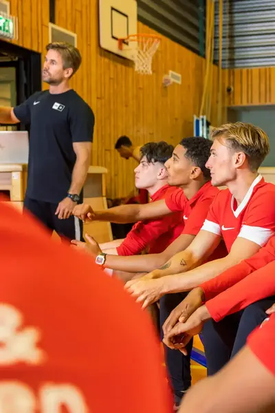 Studenten sport met rode shirts aan zitten in een gymzaal te luisteren naar de uitleg van hun docent.
