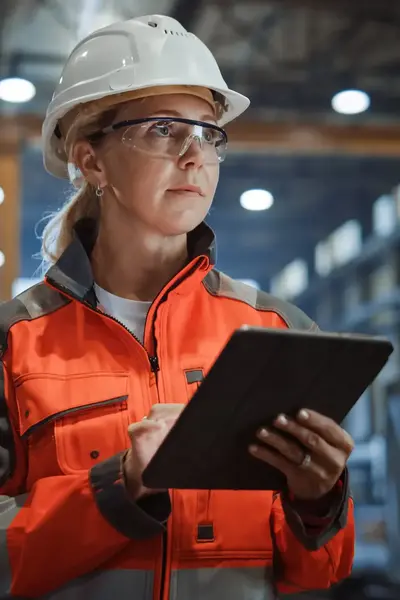 Opzichter in oranje veiligheidsjas met veiligheidsbril, helm en tablet in fabrieksetting