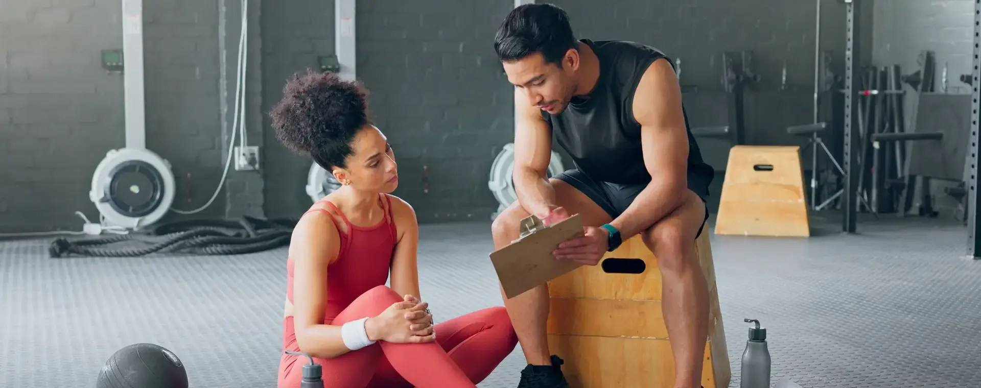 Sportinstructeur bekijkt samen met een vrouwelijke sporter een schema. Ze zitten in een gym of fintessruimte.