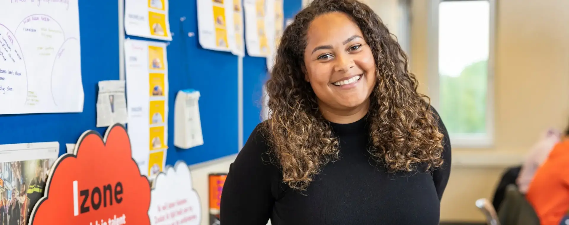 Docent met krullen glimlacht in een kleurrijke klasomgeving met educatief materiaal aan de muur.