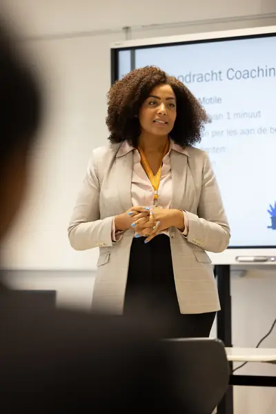 Docent geeft uitleg voor een klas en staat voor een scherm met een presentatie over coaching.