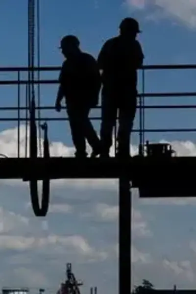 Twee personen met bouwhelmen op staan op een metalen brug bij een grote bouwplaats of industriegebied.