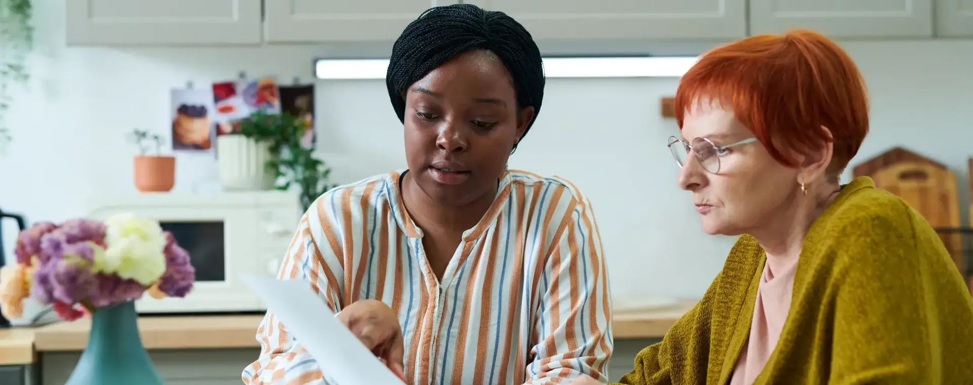 Twee vrouwen zitten aan een keukentafel en bespreken samen documenten. De jongere vrouw legt iets uit aan de oudere vrouw.