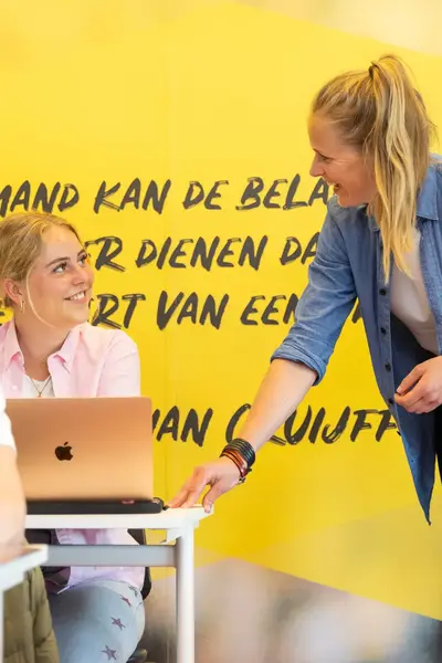 Studenten in klaslokaal met laptops en docent. Op de gele muur op de achtergrond staat een citaat van Johan Cruijff.