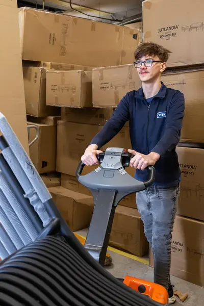 Student verplaatst een stapel stoelen met een elektrische palletwagen in een magazijn vol grote dozen.