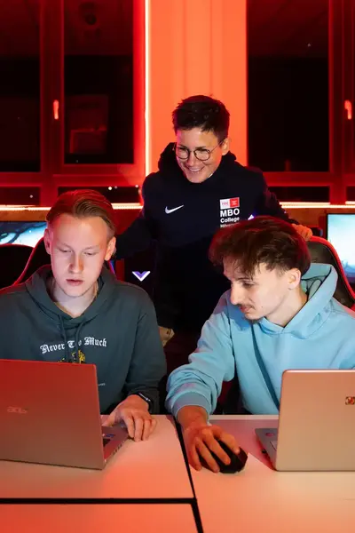 Twee studenten zitten in een speciaal game klaslokaal achter een laptop en een docent kijkt mee. Op de achtergrond zie je meerdere gamestoelen en schermen 