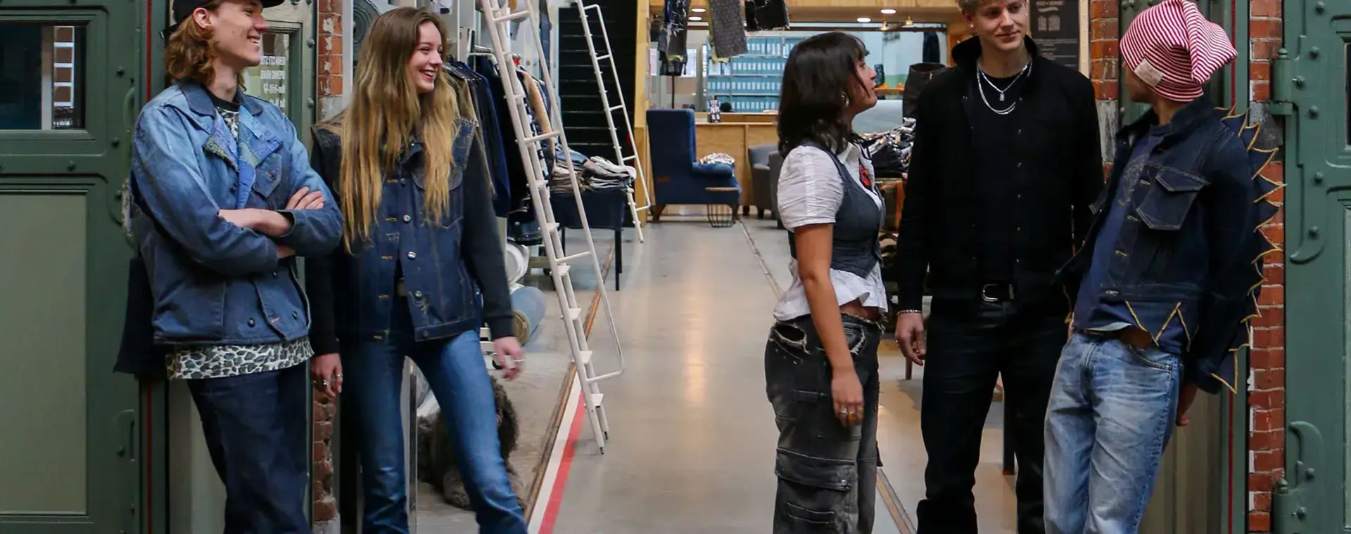 Vijf studenten in denim outfits staan samen te lachen in een creatieve modewerkplaats.