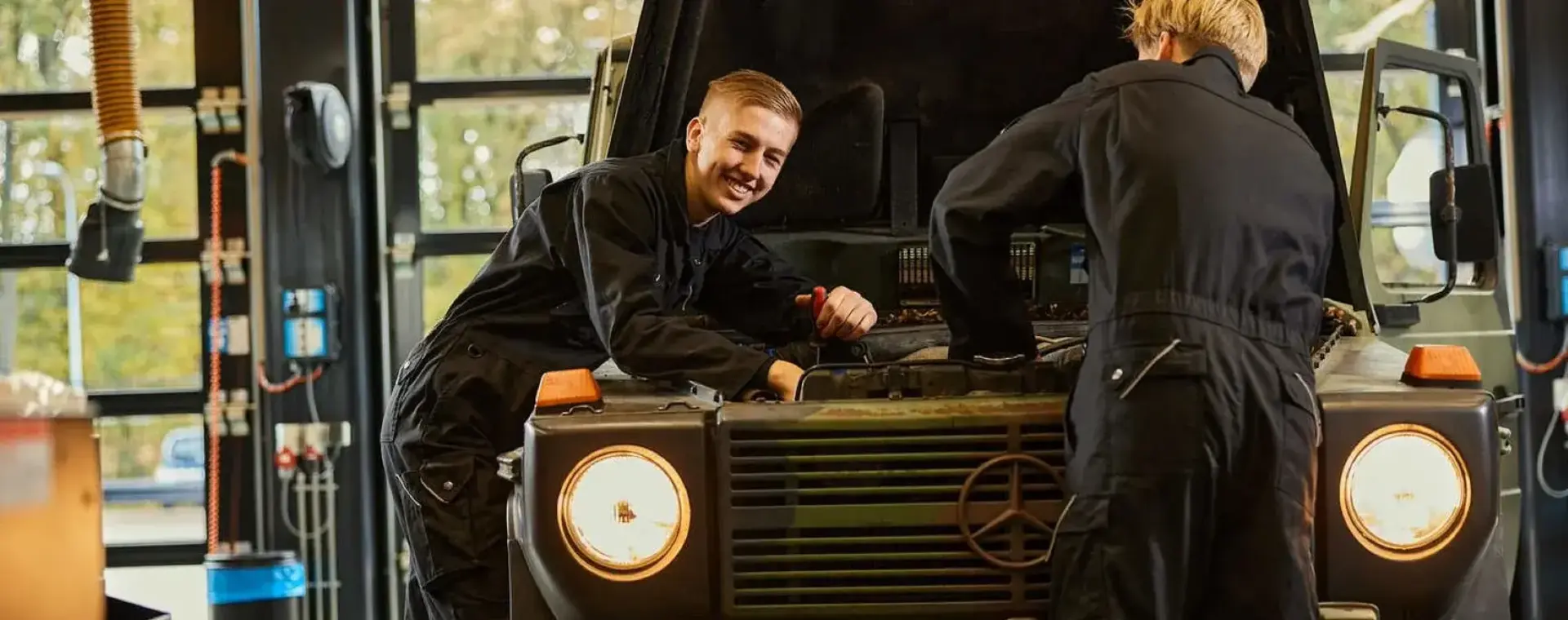 Twee studenten sleutelen aan een legervoertuig in een werkplaats, een van hen kijkt lachend naar de camera.