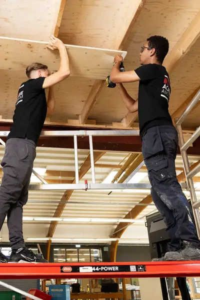 Twee studenten staan op een bouwstelling en zijn een plafond aan het maken of repareren.