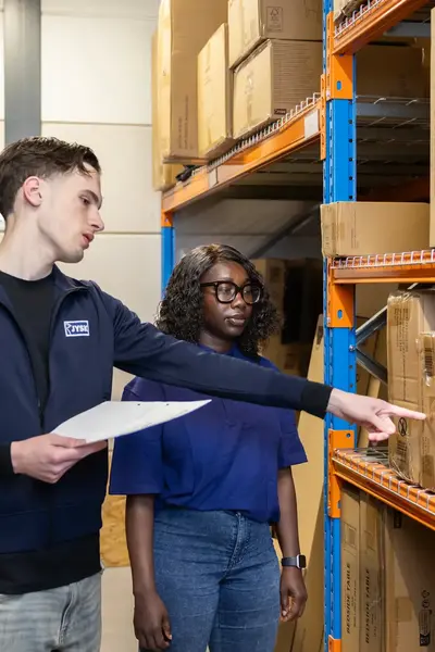 Twee studenten staan in een magazijn tussen grote dozen. Eén student wijst naar een plank terwijl de ander toekijkt.