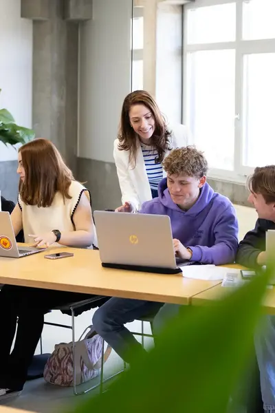 Vier studenten aan een lange tafel met geopende laptops en een docent staat achter hen en legt iets uit.