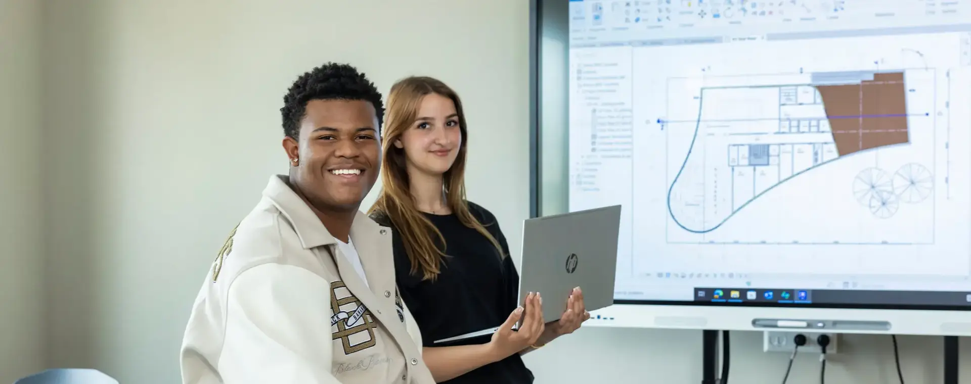 Twee studenten in klaslokaal met laptop in de hand voor een scherm met daarop een bouwtekening.