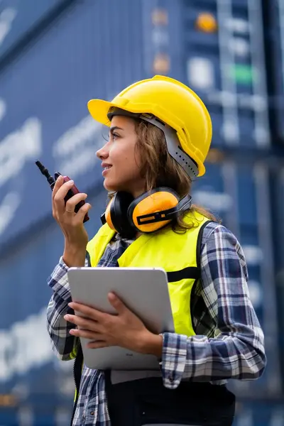 Vrouw met helm en geel hesje tussen containers geeft instructies door via een portofoon.