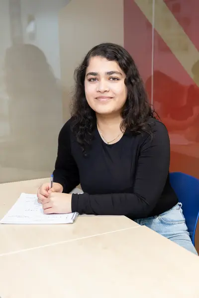 Student met donker krullend haar zit aan tafel, maakt aantekeningen in een schrift en kijkt glimlachend naar de camera.
