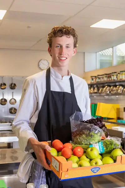 Student in koksbuis met schort toont trots een krat vol verse groenten en fruit in een keuken.