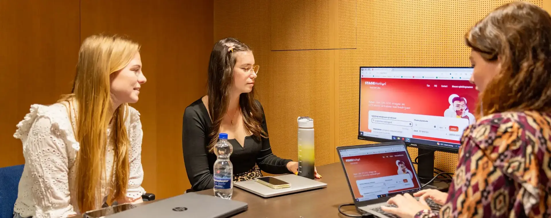 vrouw achter laptop laat info op beeldscherm aan twee jongedames zien