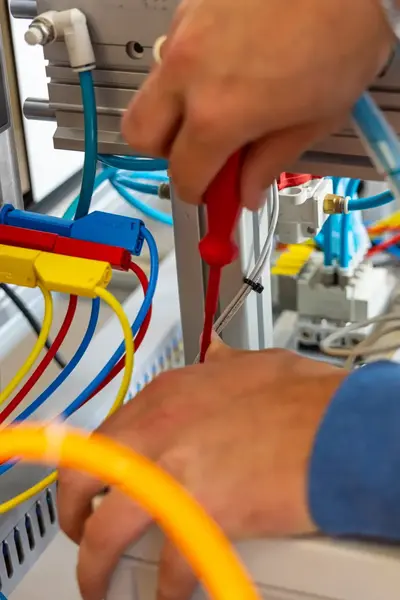 Close-up van handen die bedrading aansluiten op een besturingspaneel met gekleurde kabels en een schroevendraaier, binnen een technische installatie.