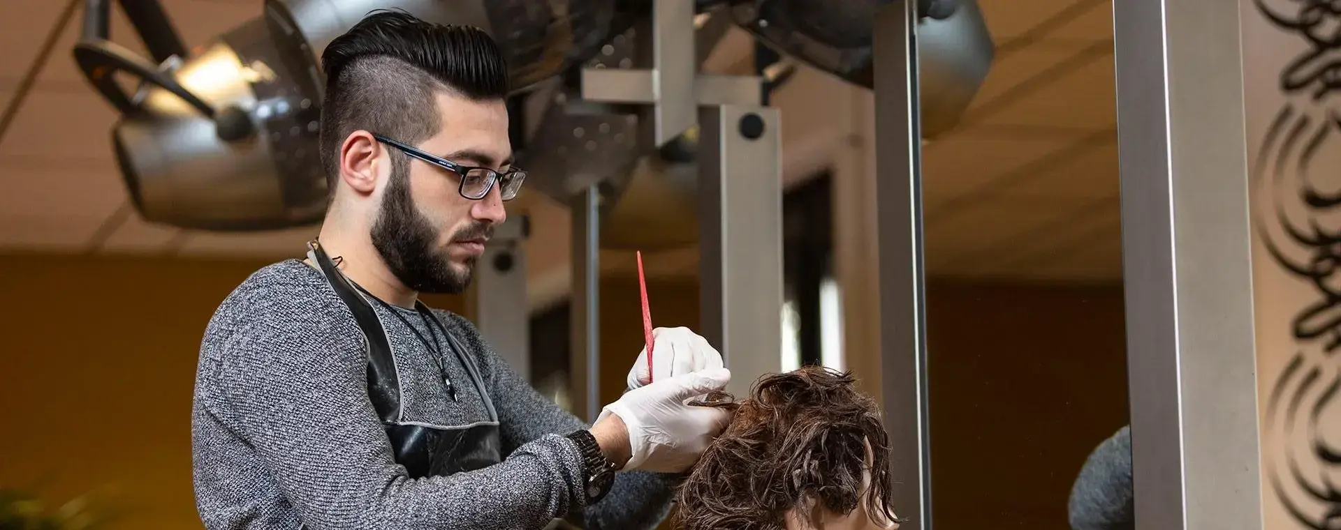 Student met handschoenen oefent het verven van haar op een oefenhoofd in een kapsalon, omringd door spiegels en droogkappen.