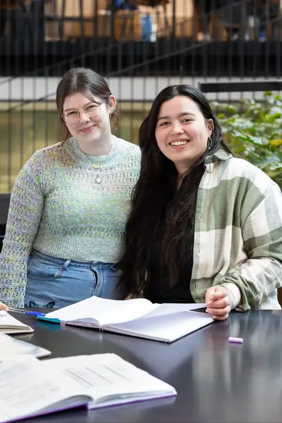 Drie studenten zitten samen aan tafel met boeken en schriften en lachen naar de camera.