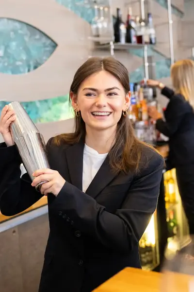 Een jonge vrouw in pak lacht achter de bar met een cocktailshaker in haar hand. Op de achtergrond maakt haar collega een drankje.