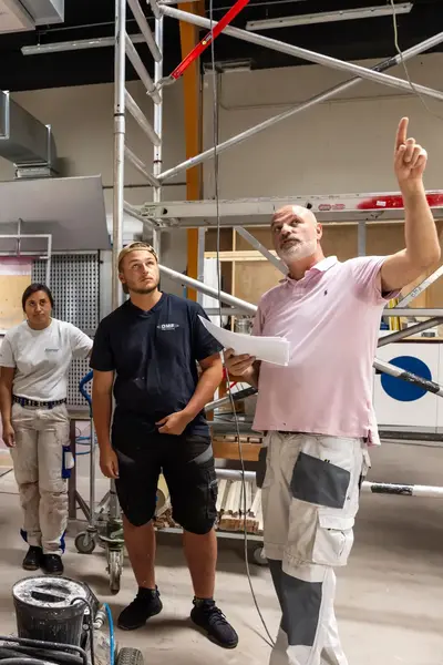 Vijf studenten en een docent staan in een praktijklokaal. De docent legt iets uit en wijst naar boven. Op de achtergrond staat een bouwsteiger.