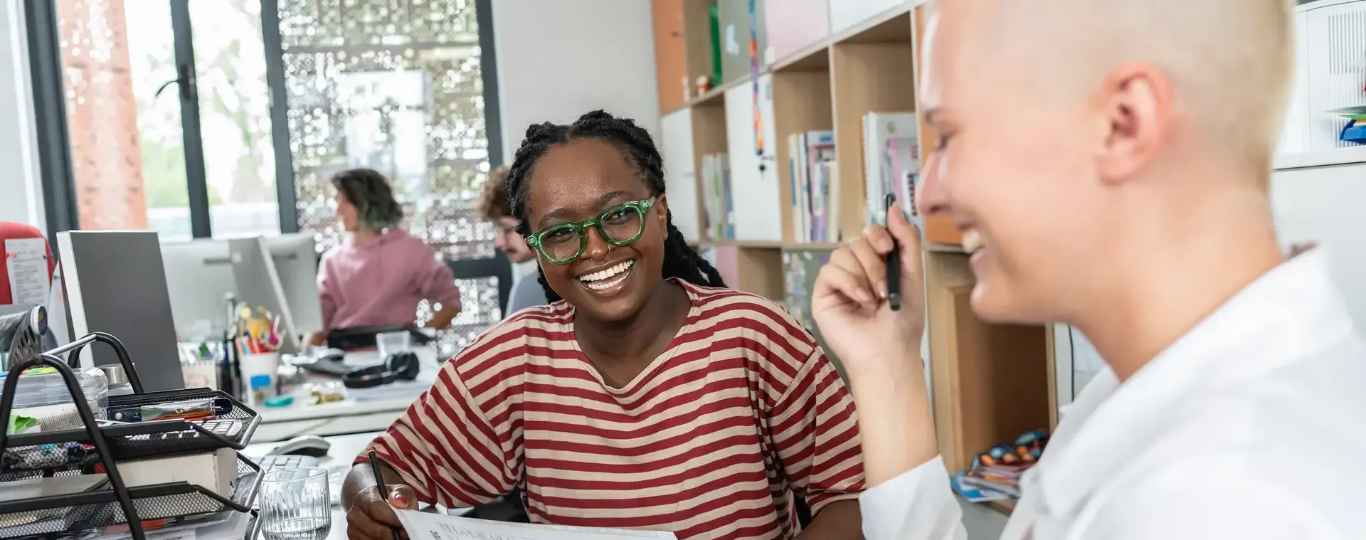 Twee vrouwen zitten samen aan een bureau in een klaslokaal of kantoorruimte. Ze lachen en overleggen terwijl één van hen een papier vasthoudt.