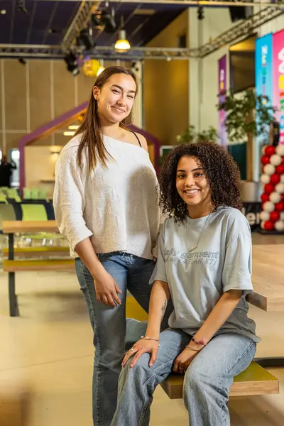 Twee studenten poseren samen in een kleurrijke en open schoolruimte met tafels en stoelen op de achtergrond.