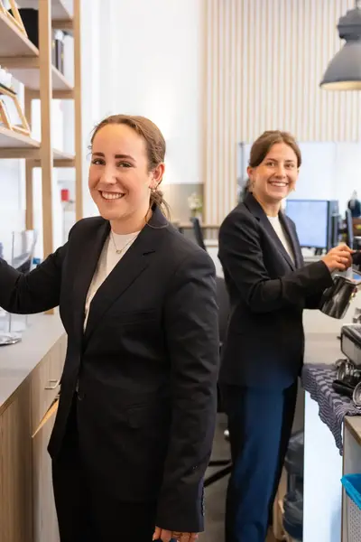 Twee vrouwen in zwarte colbertjes werken achter een koffiebar. Eén staat bij siroopflesjes, de ander maakt melk.
