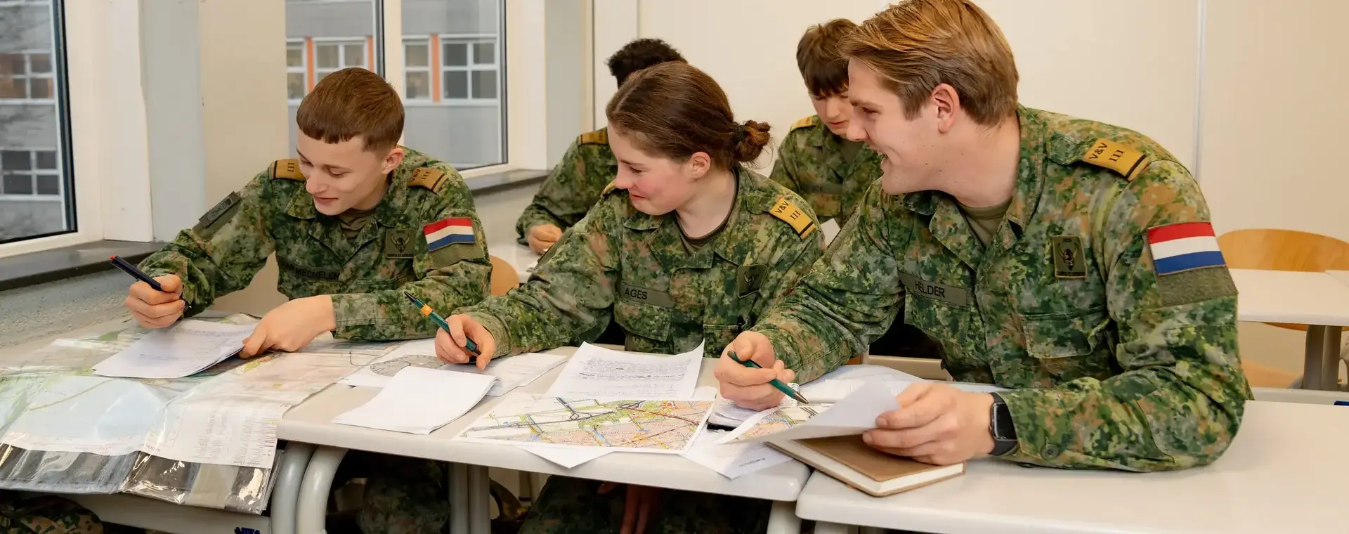 Studenten in militair uniform werken samen aan een opdracht in een klaslokaal, met kaarten en documenten op tafel.
