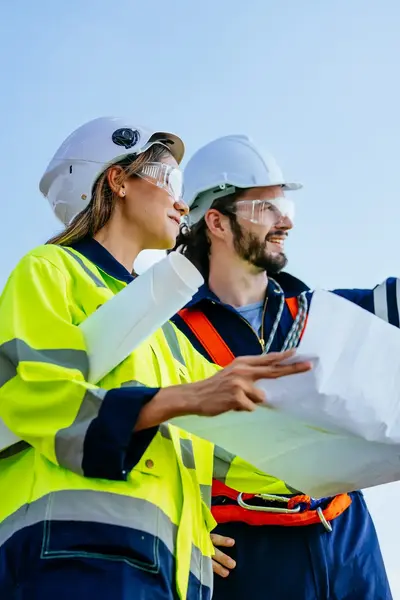 Twee collega's met helm en veiligheidsbril bestuderen bouwtekening bij windmolen
