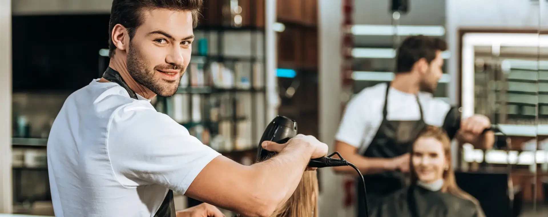 Student in opleiding tot kapper föhnt het haar van een klant in een moderne salon, terwijl hij glimlachend in de camera kijkt. In de spiegel zie je de klant met een tevreden glimlach.